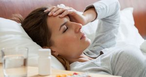 A woman is lying in bed with a headache, on her nightstand are some medications